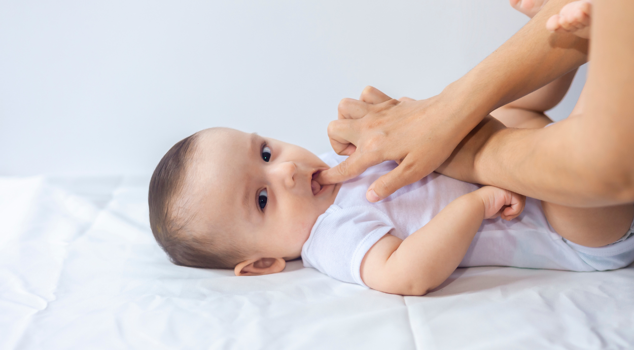 Baby lying on a white sheet, playfully sucking on an adult's finger.