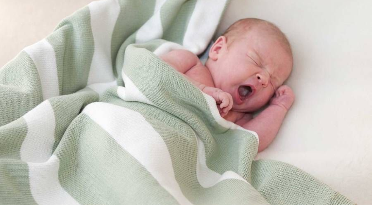 A close-up of a cozy, swaddled baby resting under a soft, green and white striped blanket. The scene evokes warmth and tenderness.