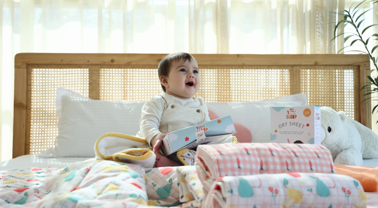 A baby sits on a bed surrounded by colorful blankets and plush toys, with educational sleep products on display. Sunlight filters through curtains.