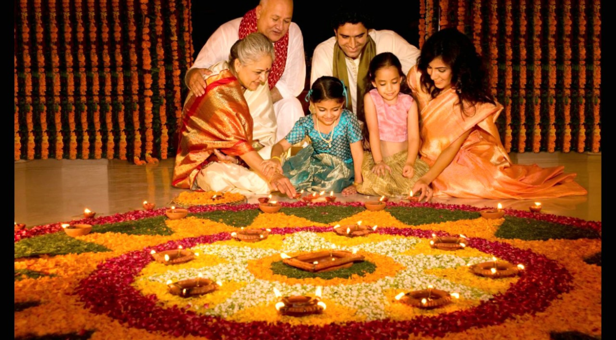 A family gathers around a colorful flower rangoli, decorated with candles, celebrating a festive occasion. Traditional attire is visible.