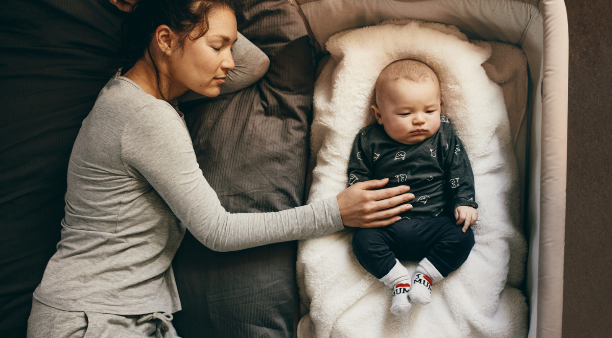 A mother gently rests her hand on her baby, who is lying on a soft surface in a cozy setting.