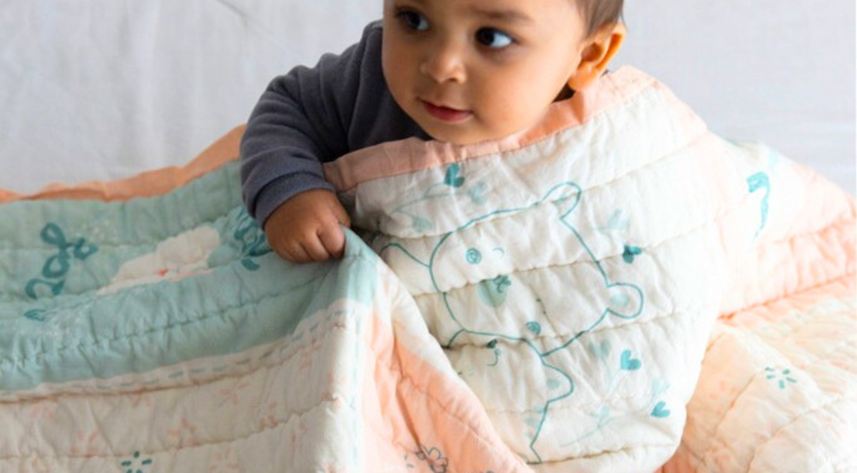 A child wrapped in a pastel-colored quilt featuring cute illustrations, sitting against a soft background.