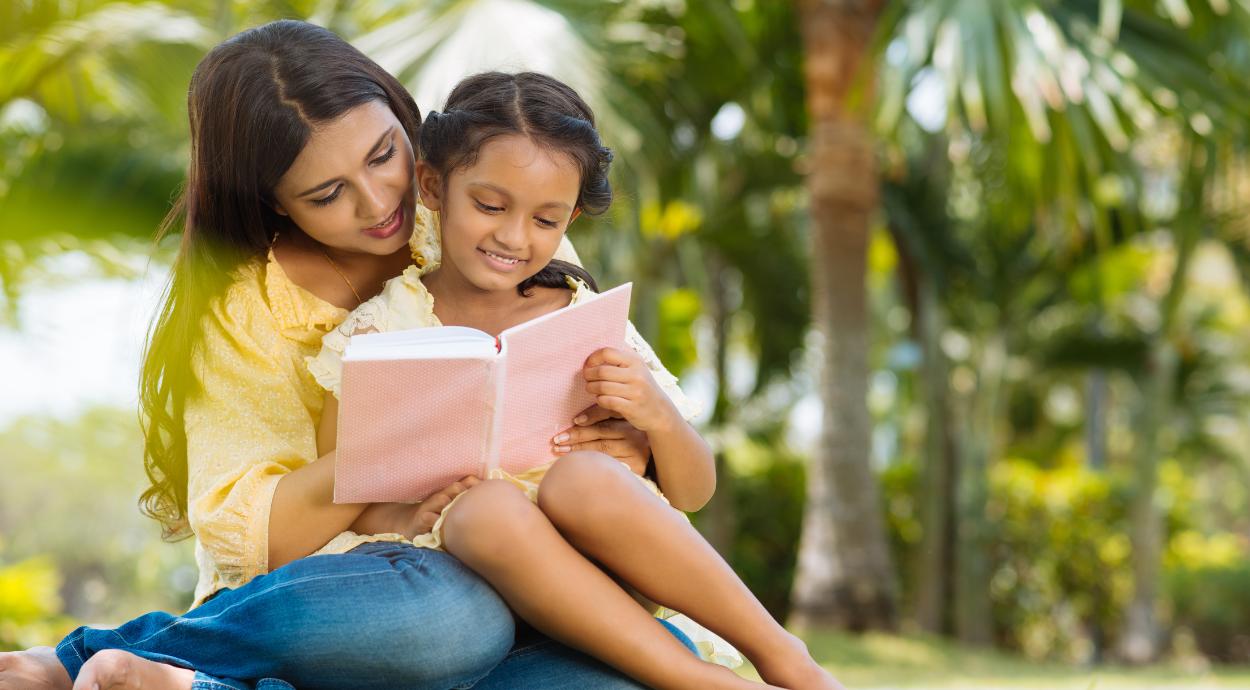A mother and her young daughter sit together outdoors, reading a book in a lush, green setting with palm trees in the background