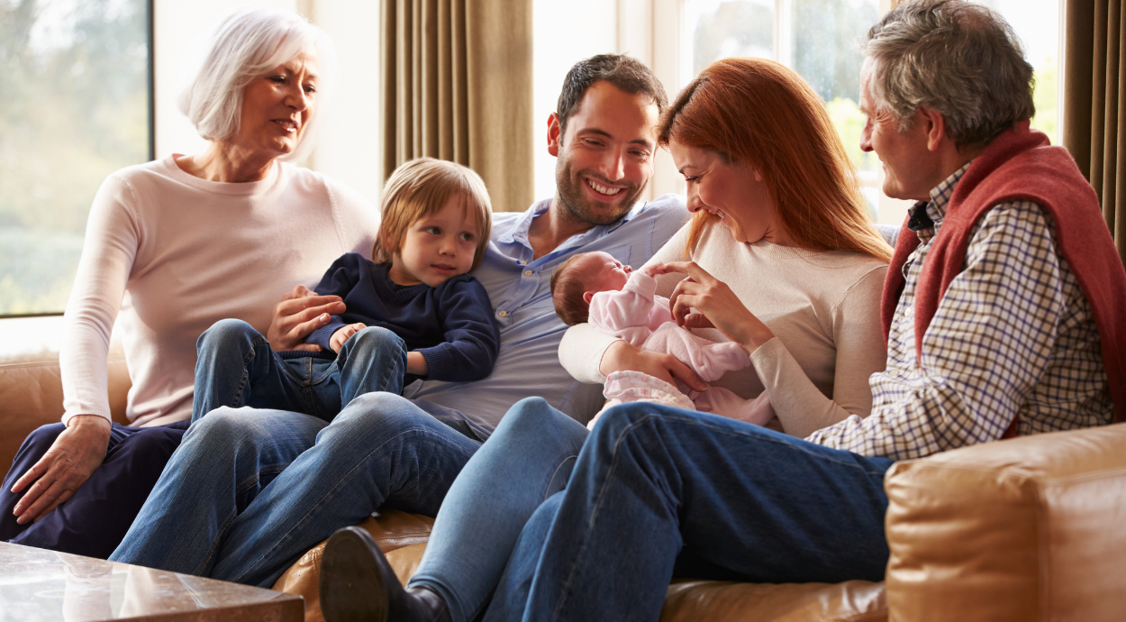 A family sits together on a couch, with three adults and three children, sharing a cozy moment in a sunlit room.