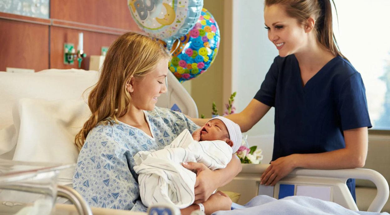 A mother in a hospital gown gently holds her newborn in a cozy hospital room, while a nurse offers support and encouragement.