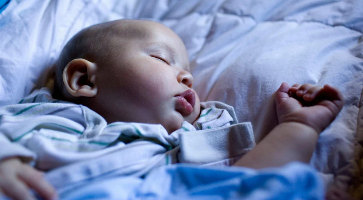 Sleeping baby lying on a white bed, wearing a striped outfit, with one arm stretched out and relaxed.
