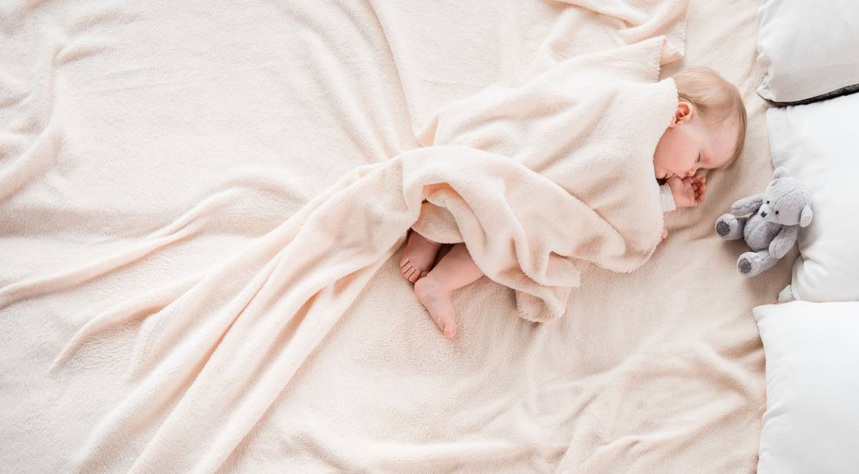 A cozy scene of a child wrapped in a soft, pale blanket, with a plush toy nearby, resting on a fluffy blanket surrounded by pillows.