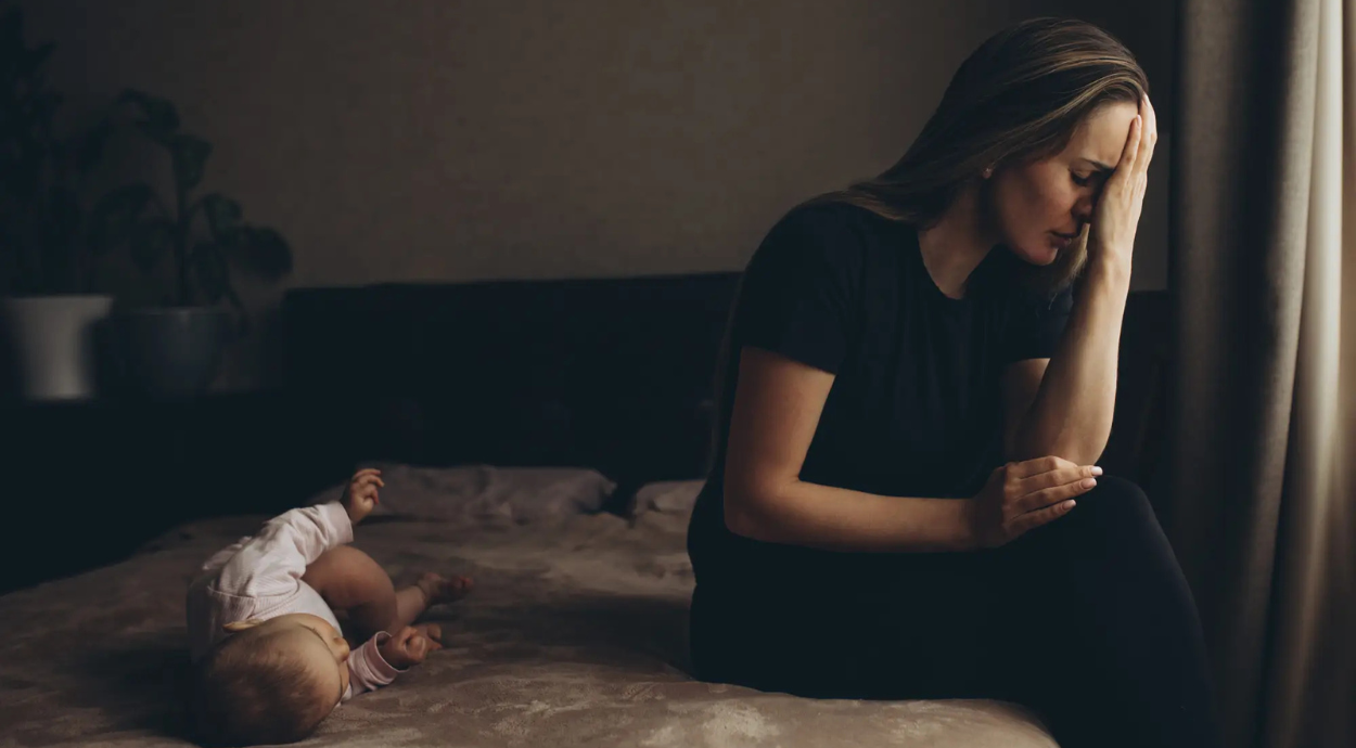 A woman sits pensively on a bed, while a baby lies playfully nearby, captured in soft, warm lighting.