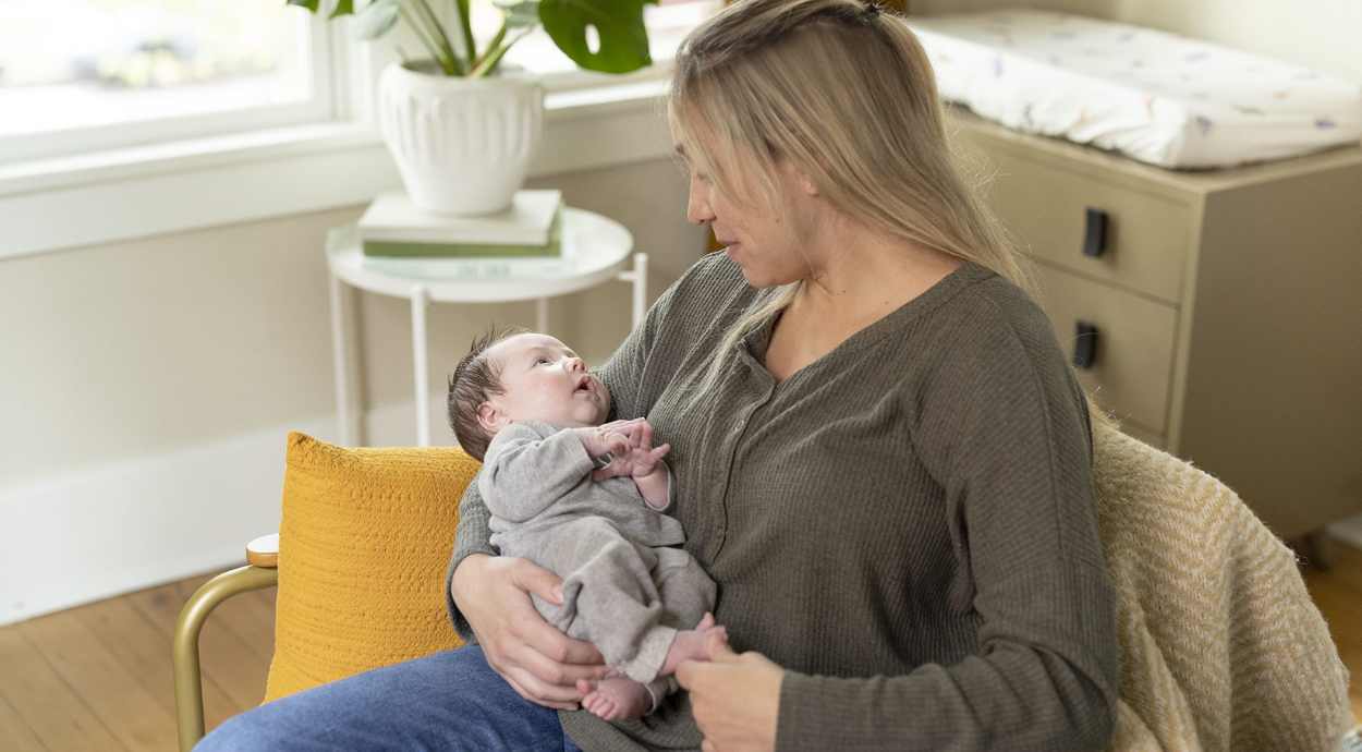 A mom gently holds a dressed baby in a cozy living room, surrounded by soft furnishings and natural light.
