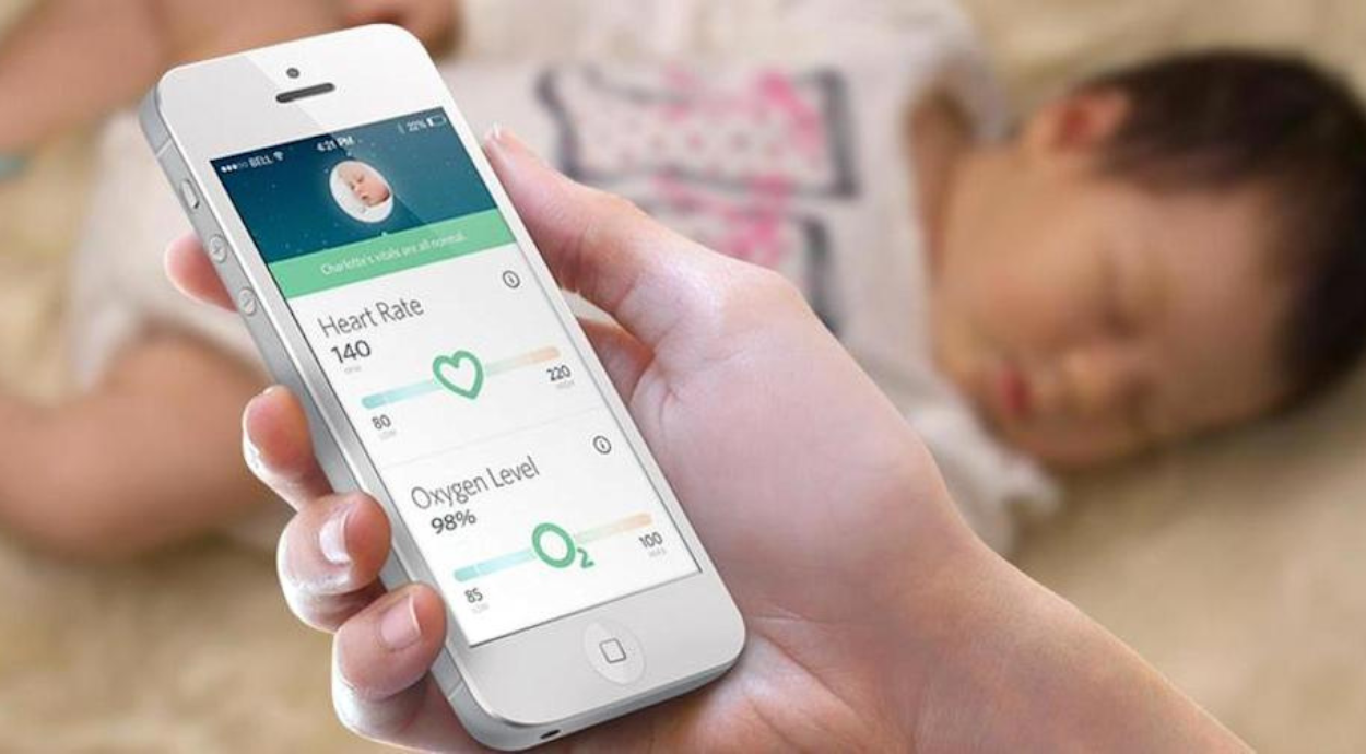 A hand holding a smartphone displays a health app showing heart rate and oxygen level of a sleeping baby.