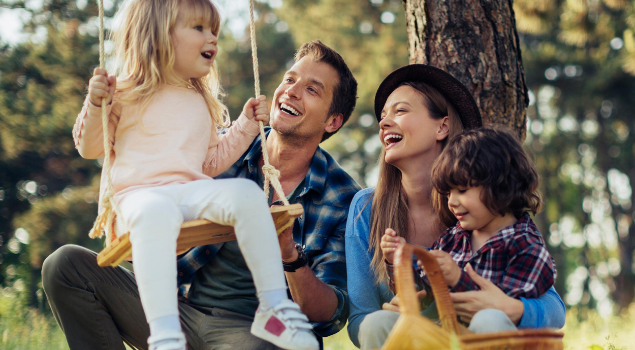 A family enjoys a sunny day outdoors, with two children playing on swings and parents nearby, surrounded by trees and nature.