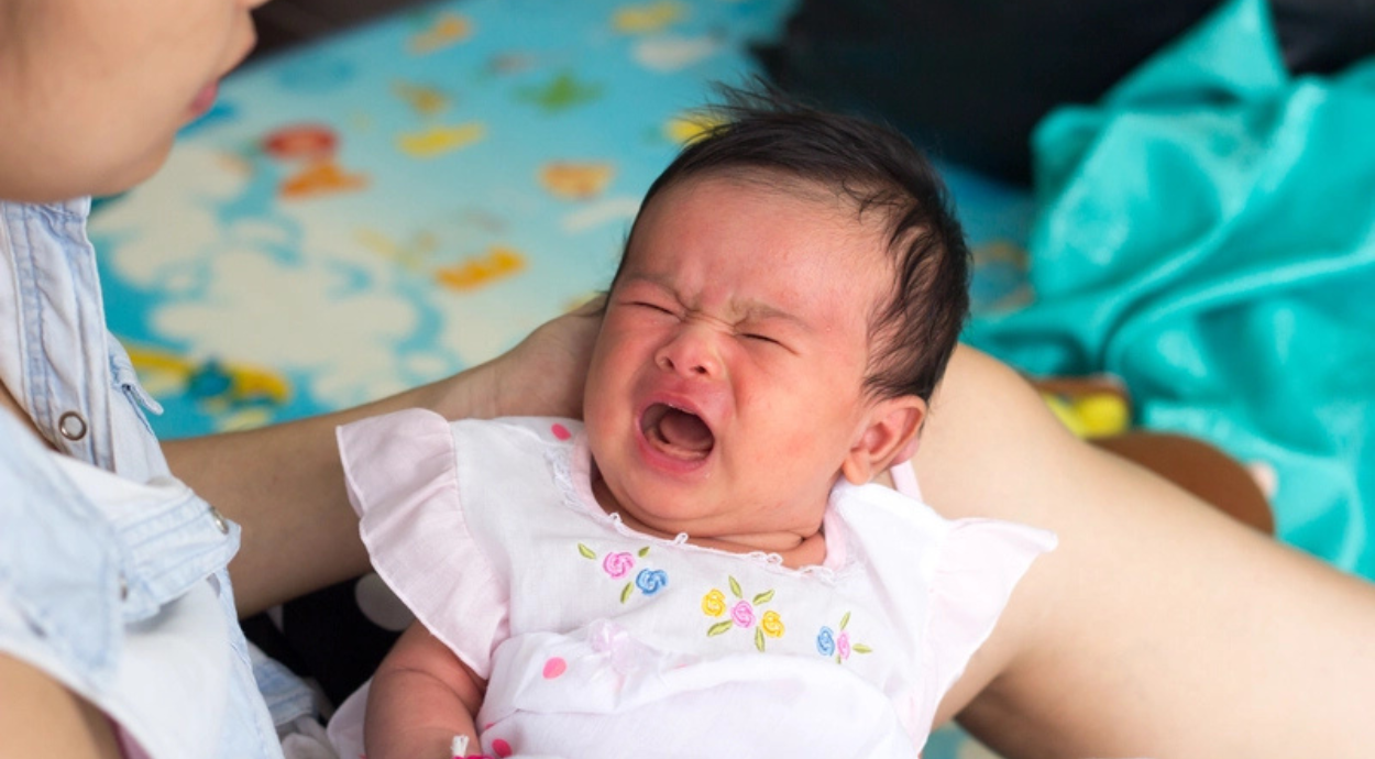 A person holds a baby wearing a pink dress with colorful embroidery, sitting on a vibrant, patterned play mat.