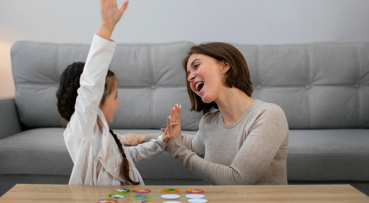 A woman and a young girl joyfully clap hands at a table with colorful circular pieces. The gray sofa in the background adds a cozy, cheerful atmosphere.