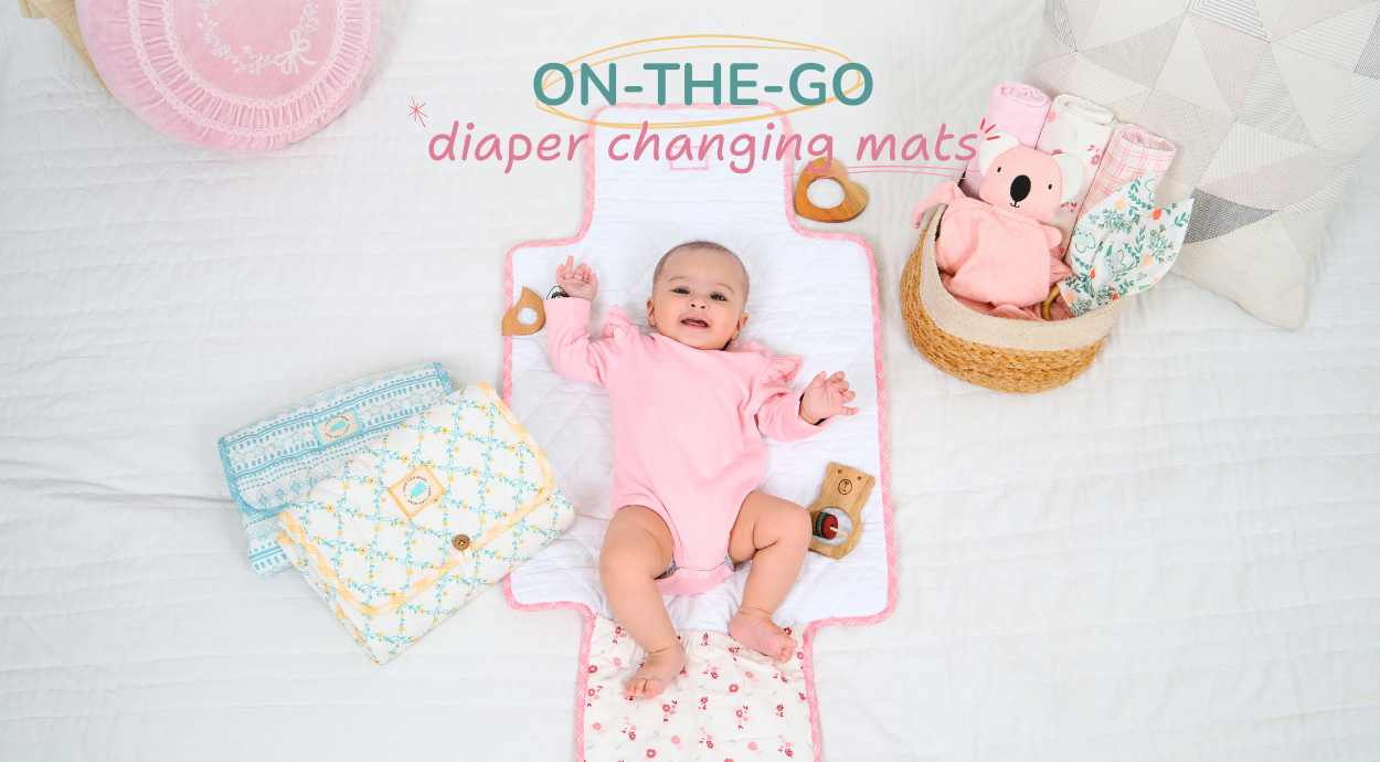 A baby in a pink onesie lies on a diaper changing mat, surrounded by colorful baby clothes and a plush toy in a woven basket.
