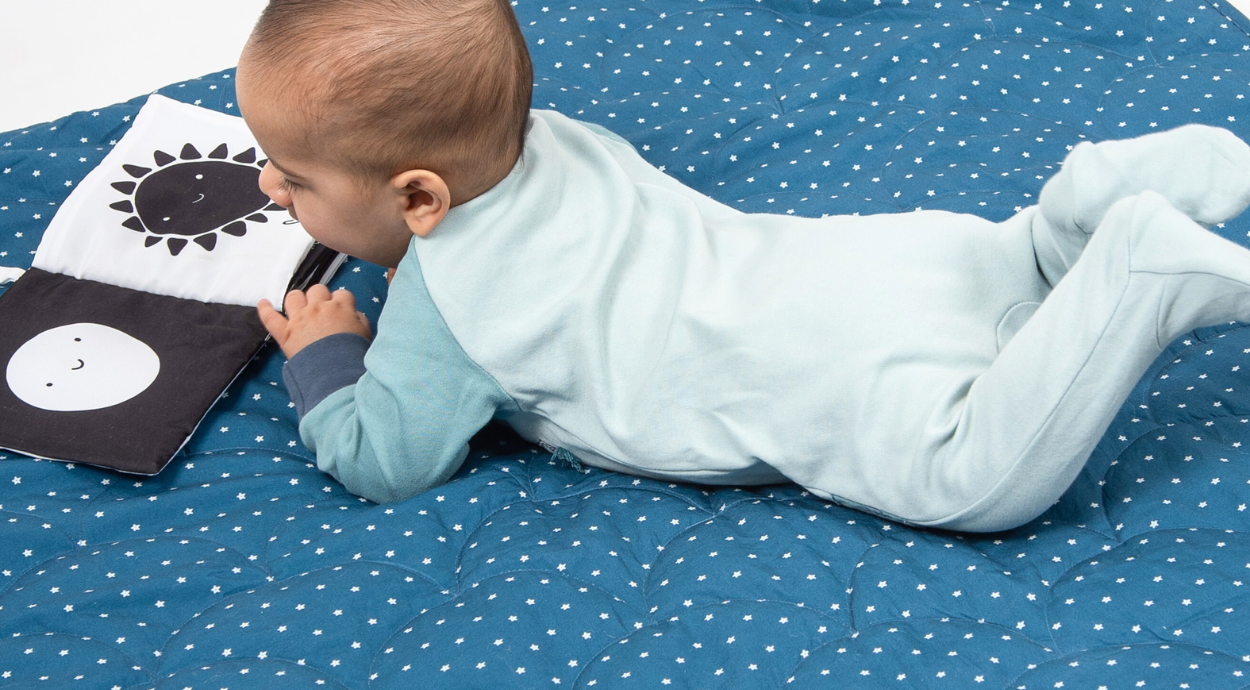 A baby in a light blue onesie lies on a patterned blue messy mat, engaging with a black and white cloth book.