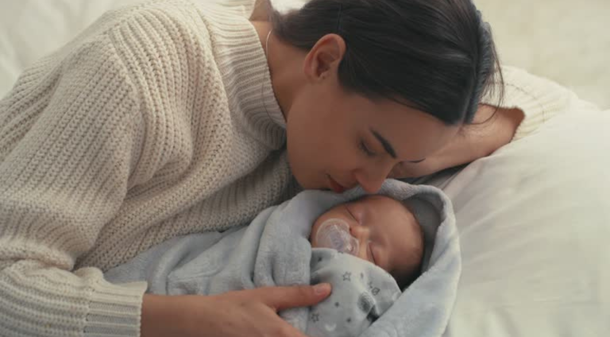 A woman in a cozy sweater lovingly embraces a sleeping baby swaddled in a grey blanket. The scene is serene and intimate, conveying warmth and tenderness.