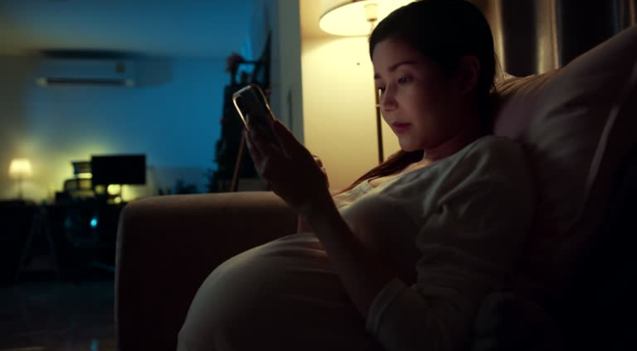 Pregnant woman in a cozy dimly-lit room, sitting on a sofa, absorbed in her phone. Soft lamp glow casts a serene and reflective ambiance.