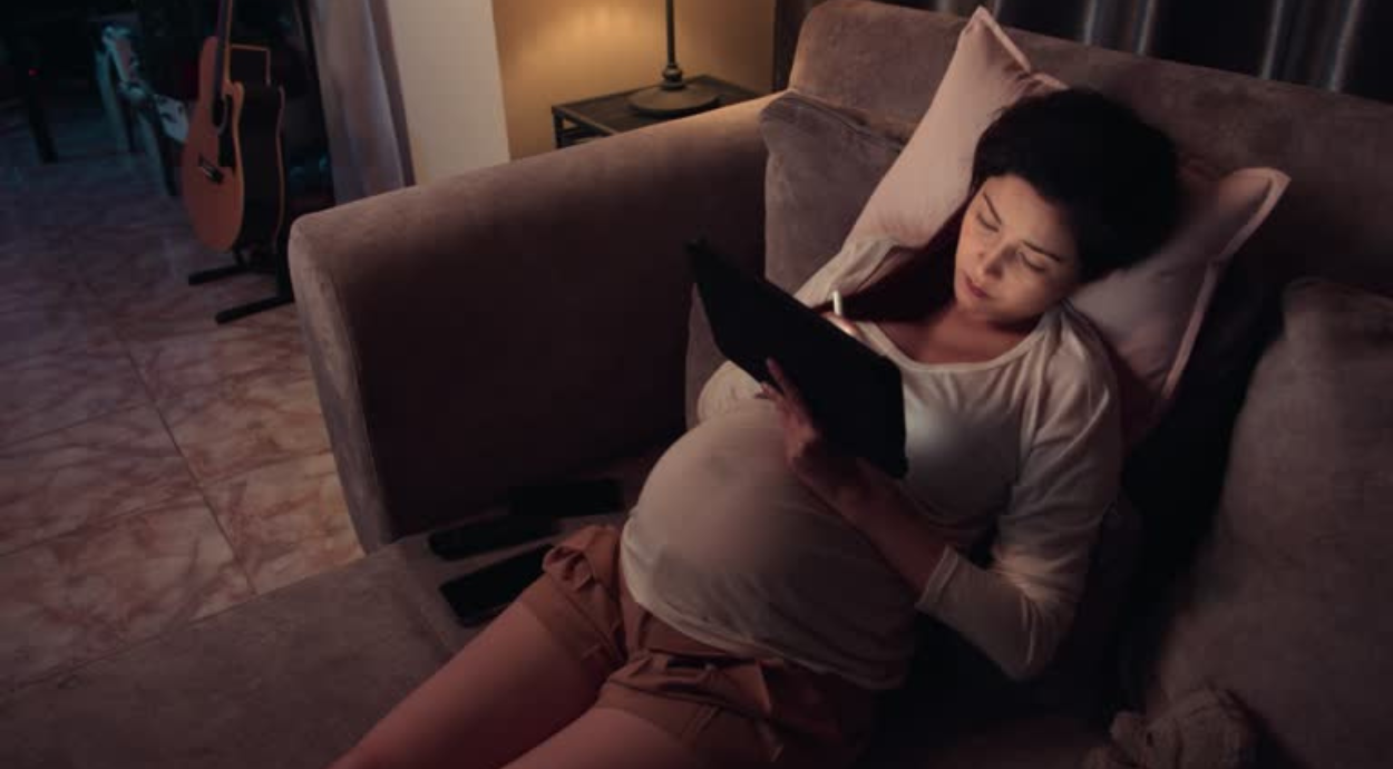 A pregnant woman is lying on a couch, using a tablet with a stylus. The setting is softly lit, creating a calm and focused atmosphere. A guitar is visible in the background.