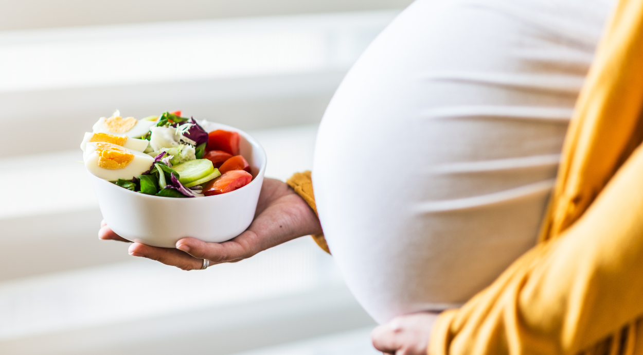 Pregnant woman holding a bowl of fresh salad with eggs, cucumbers, tomatoes, and greens.