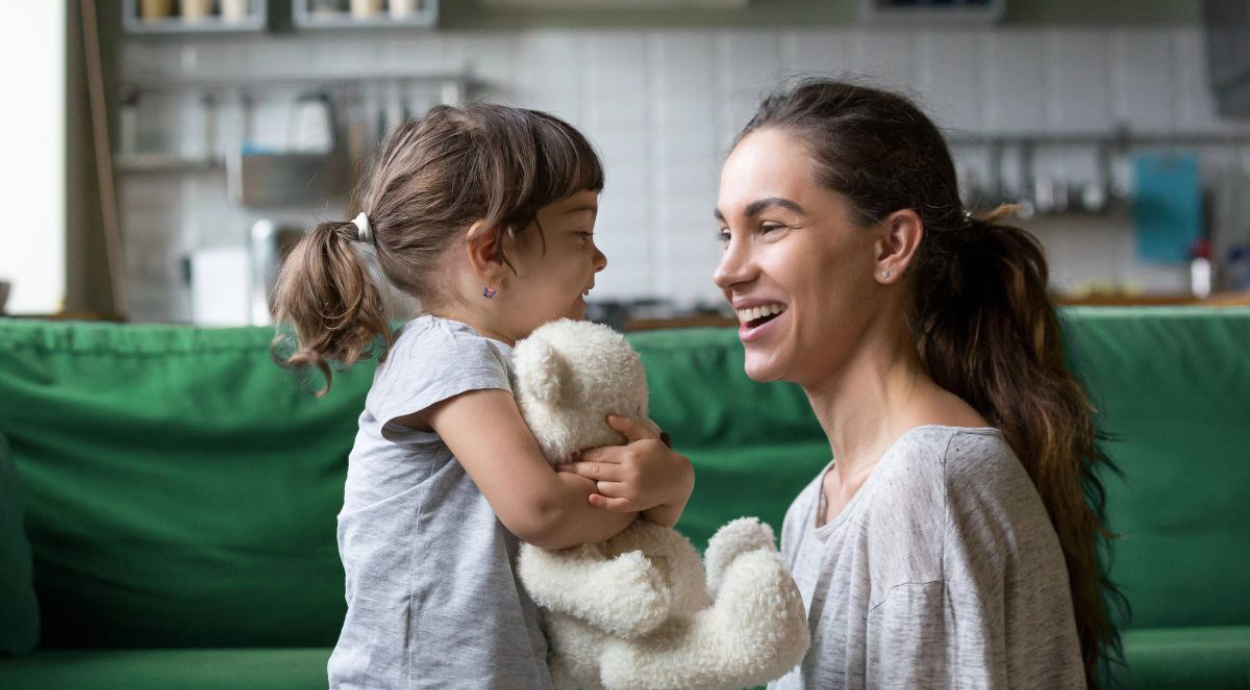 A young girl in a gray shirt, holding a plush bear, stands closely to a woman in a gray top on a green couch at home.