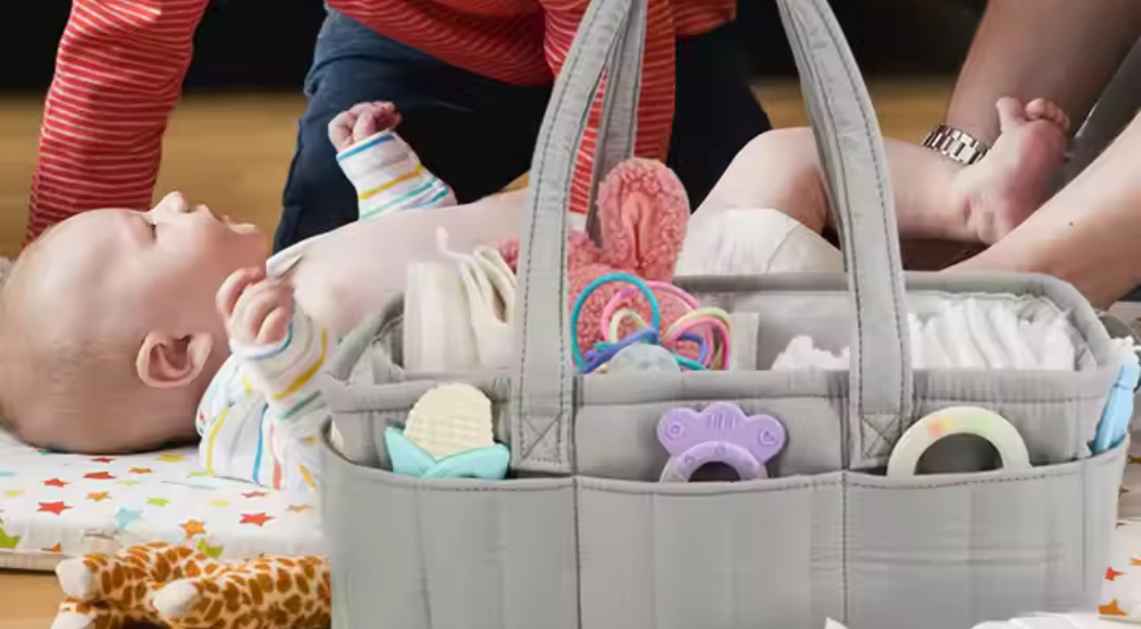 A baby lies on a colorful play mat near a gray diaper bag filled with toys and supplies, while a caregiver attends to them.