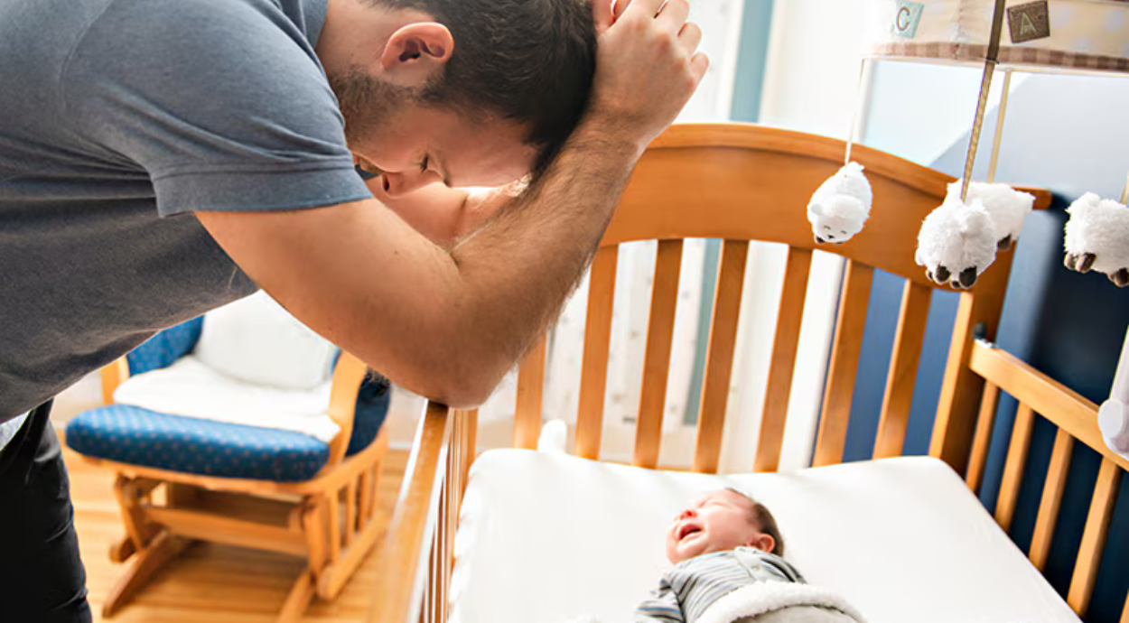 A tired parent leans over a crib, gazing at their sleeping baby, surrounded by a cozy nursery with a rocking chair and soft toys.