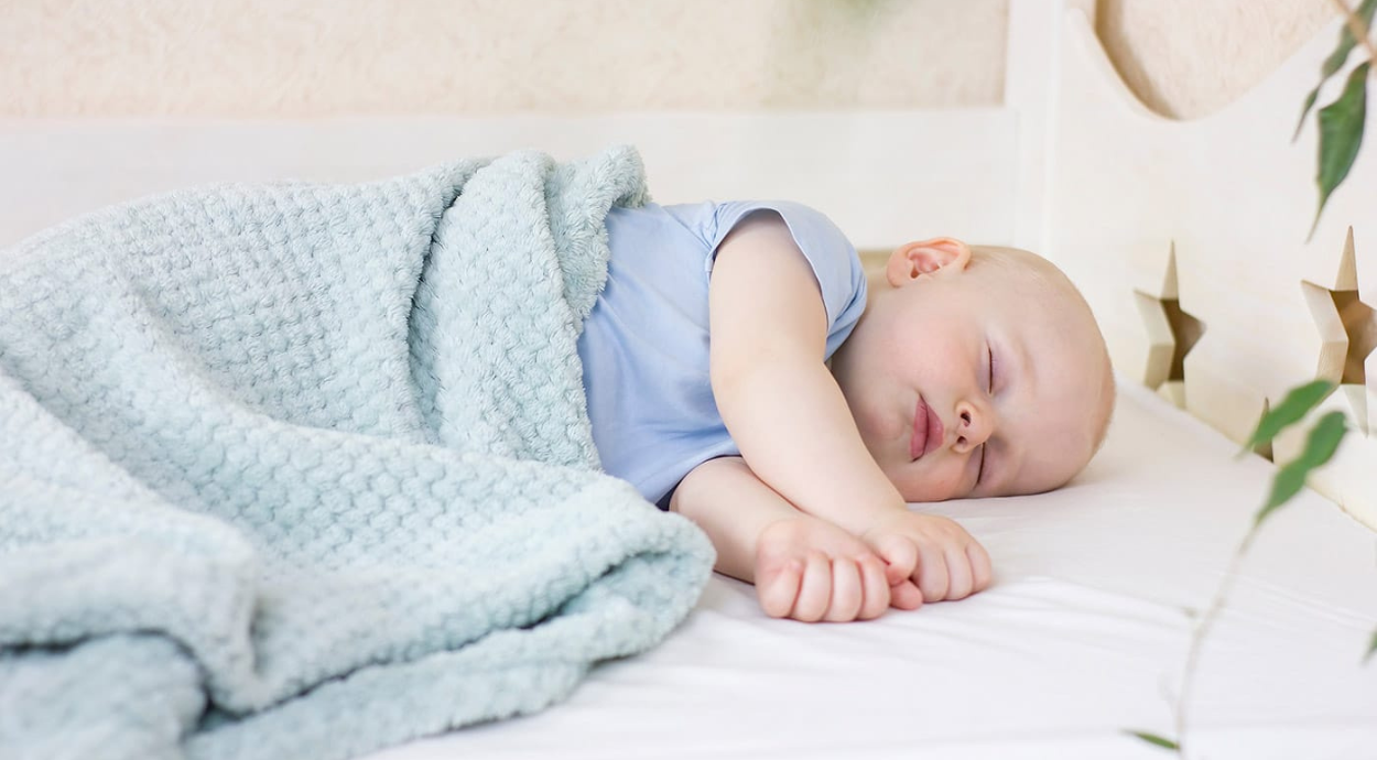 Baby sleeping peacefully on a bed, covered with a soft light blue blanket, in a cozy, softly lit room.