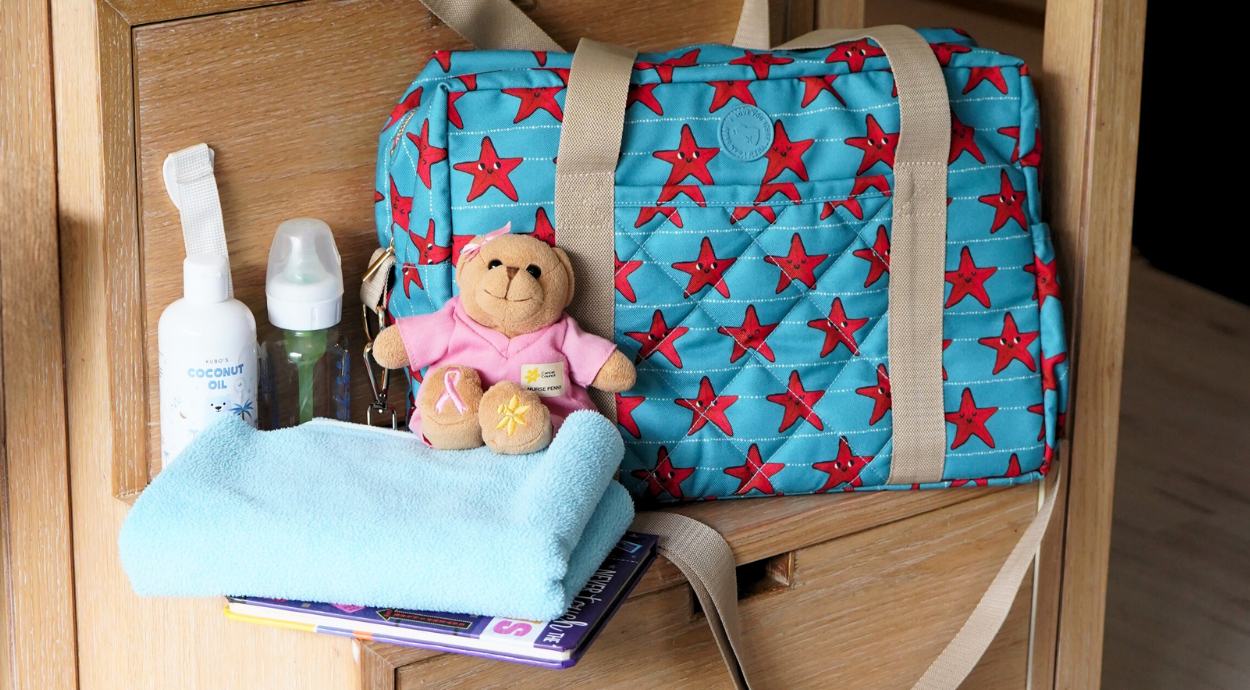 A colorful diaper bag with red stars rests on wooden stairs, surrounded by baby essentials: lotion, towel, plush teddy bear, and baby toy, creating a warm, organized scene.