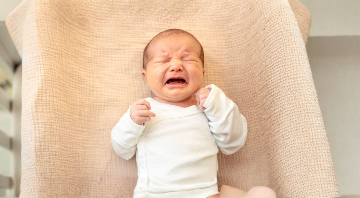 A baby dressed in a white outfit sits on a soft, textured blanket, looking curiously at the camera.