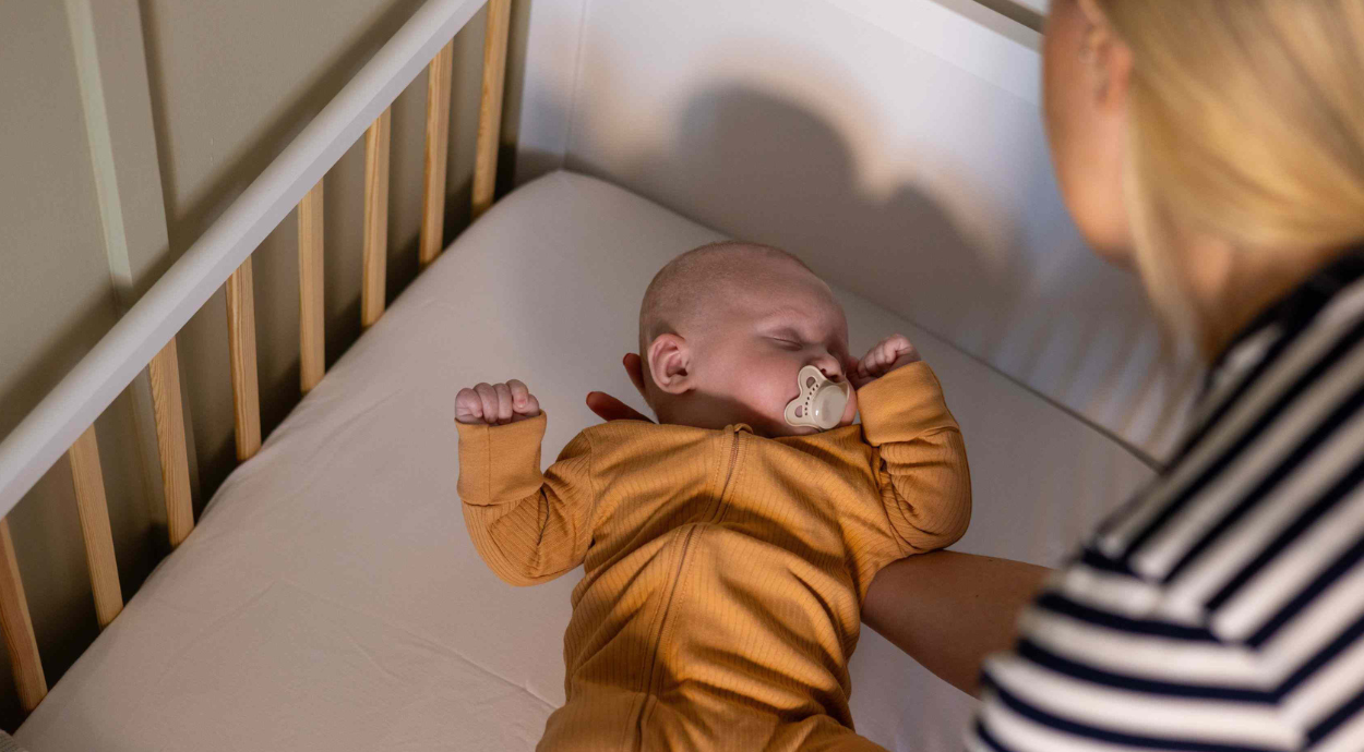 A baby in a cozy, brown outfit lies in a crib, with a caregiver nearby, creating a warm and nurturing atmosphere.