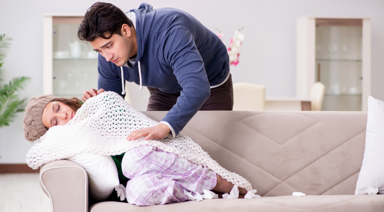 A man tenderly drapes a blanket over a woman curled up on a beige sofa, creating a cozy, nurturing atmosphere at home.