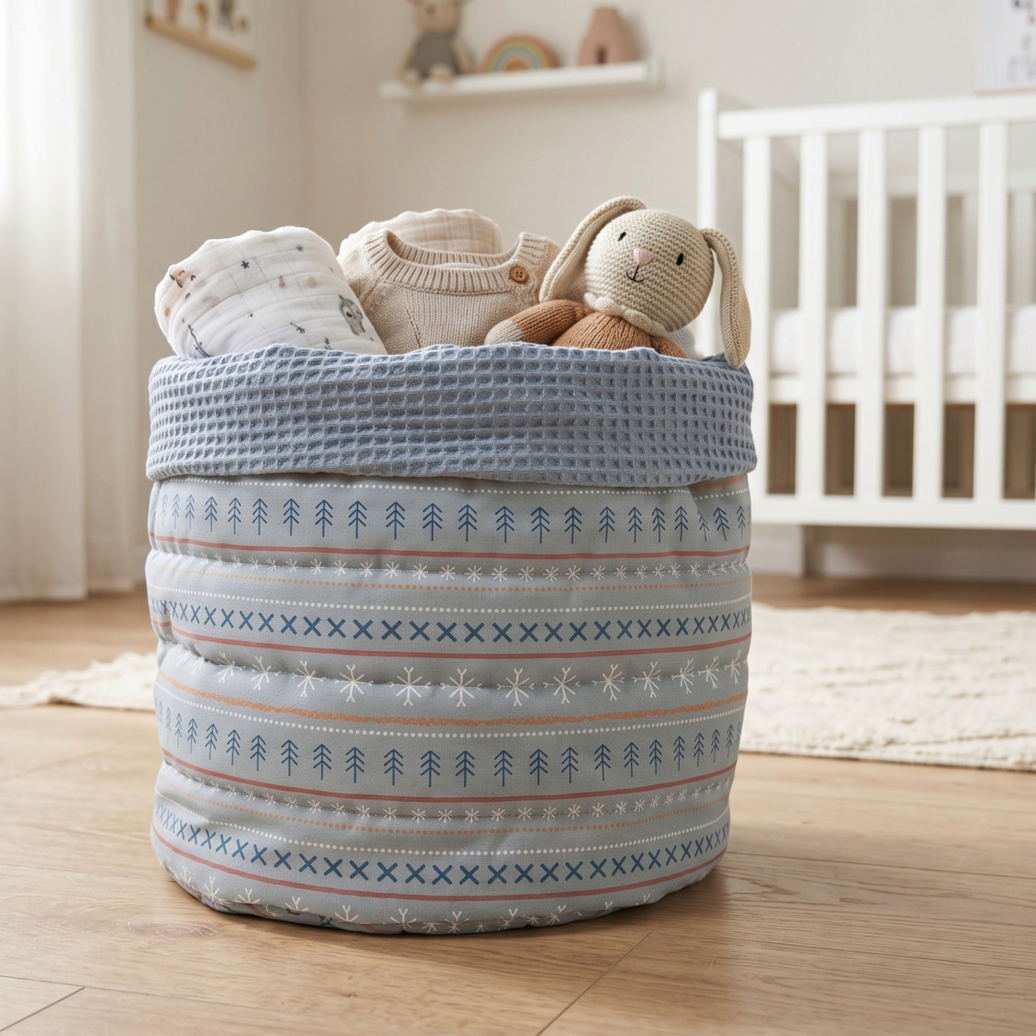 A round fabric storage basket with a Nordic tree and snowflake pattern in blue, beige, and white, filled with baby clothes and a knitted bunny toy in a softly lit nursery.