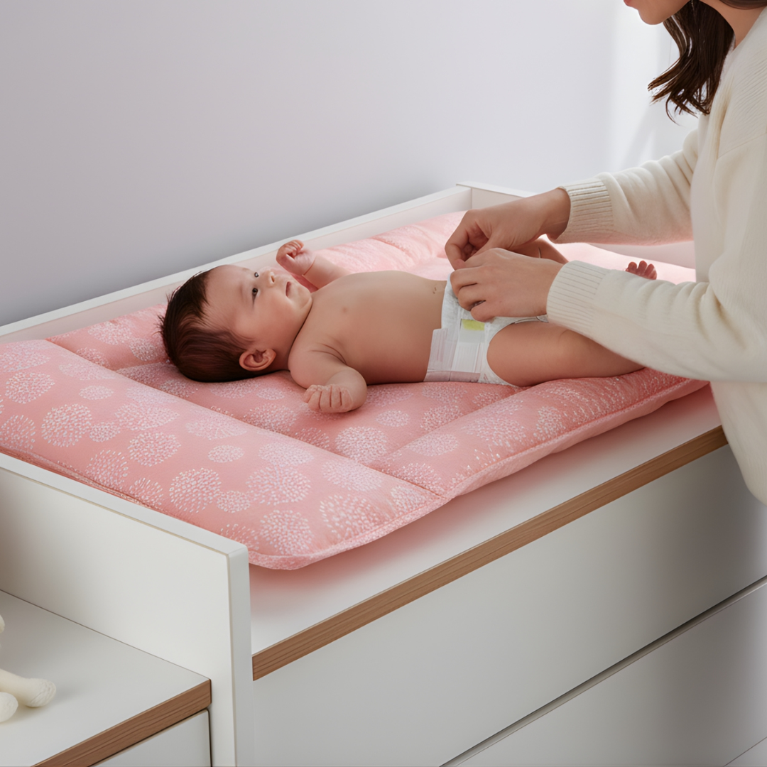 A baby lying on a pink patterned changing station on a white changing table, while an adult adjusts the baby's diaper.