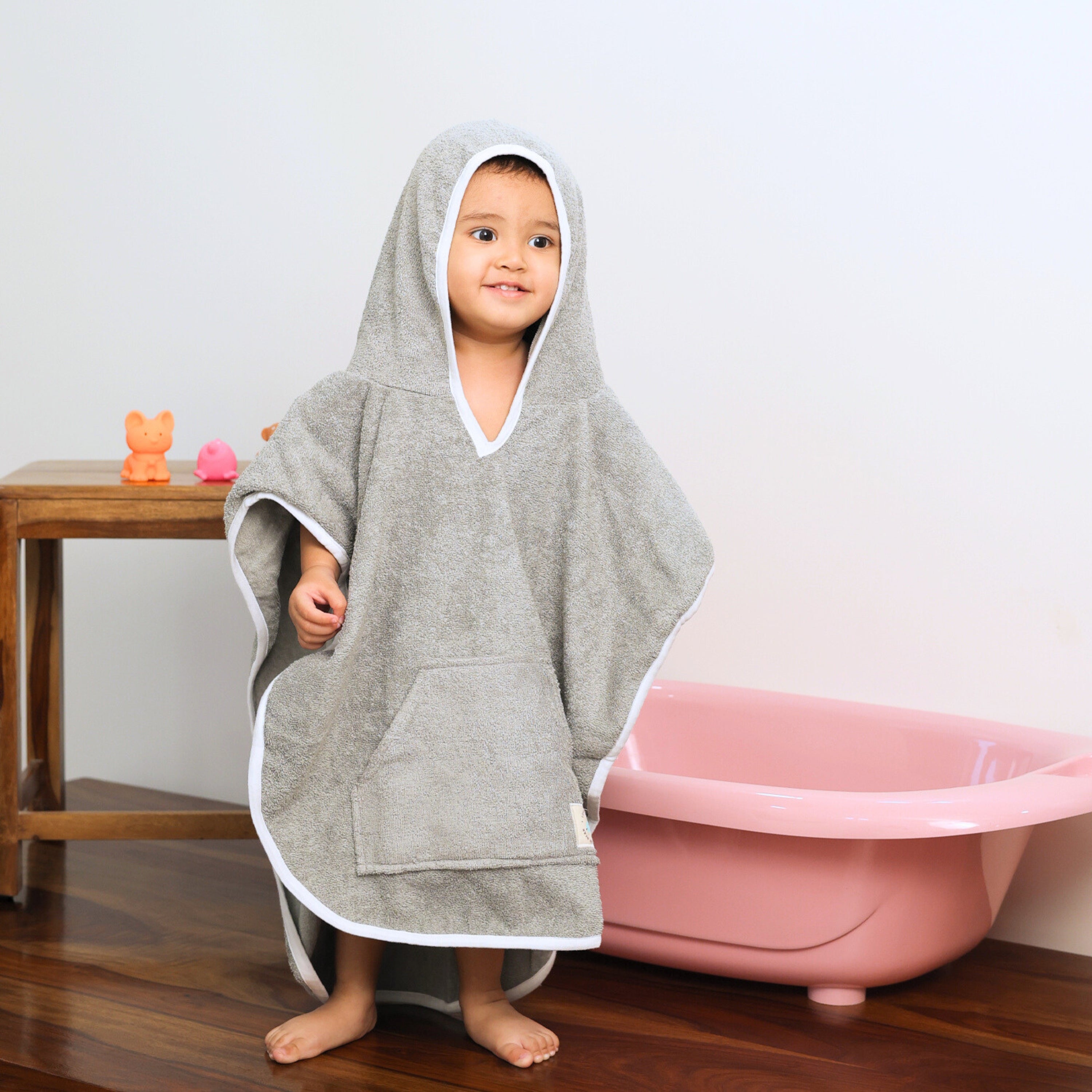 A toddler standing indoors wearing a gray hooded towel with white trim and a front pocket. A pink bathtub and wooden table with toy animals are visible in the background.