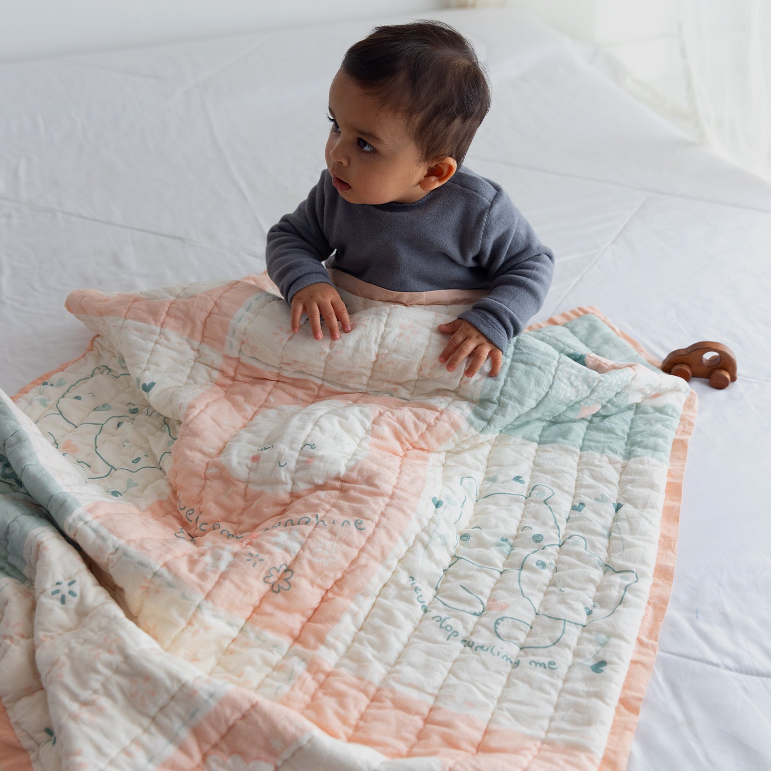 A baby wrapped in a pastel quilt adorned with cute animal illustrations, relaxing on a light-colored bed. A wooden toy is nearby.
