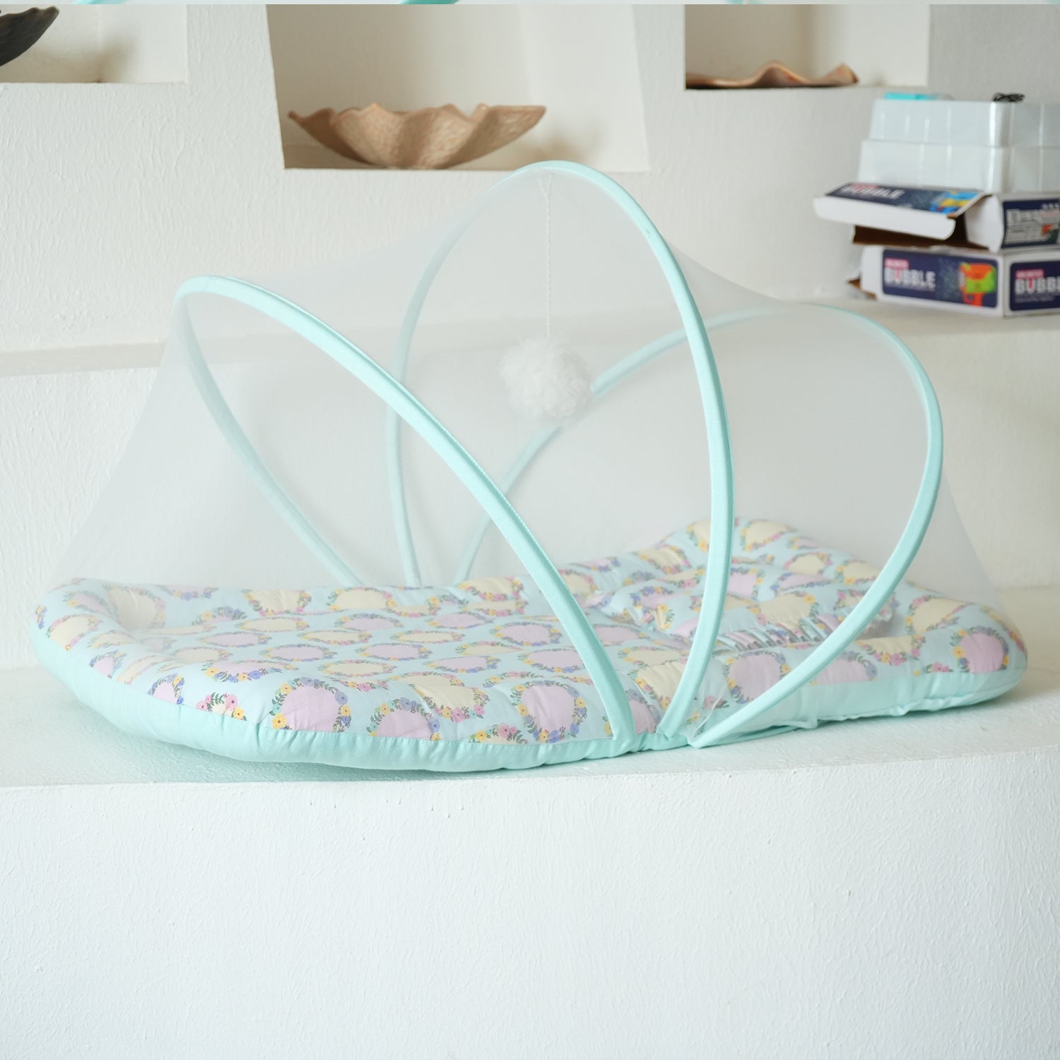 A soft, pastel-colored baby gadda set with a mosquito net, featuring a floral pattern and a hanging pom-pom