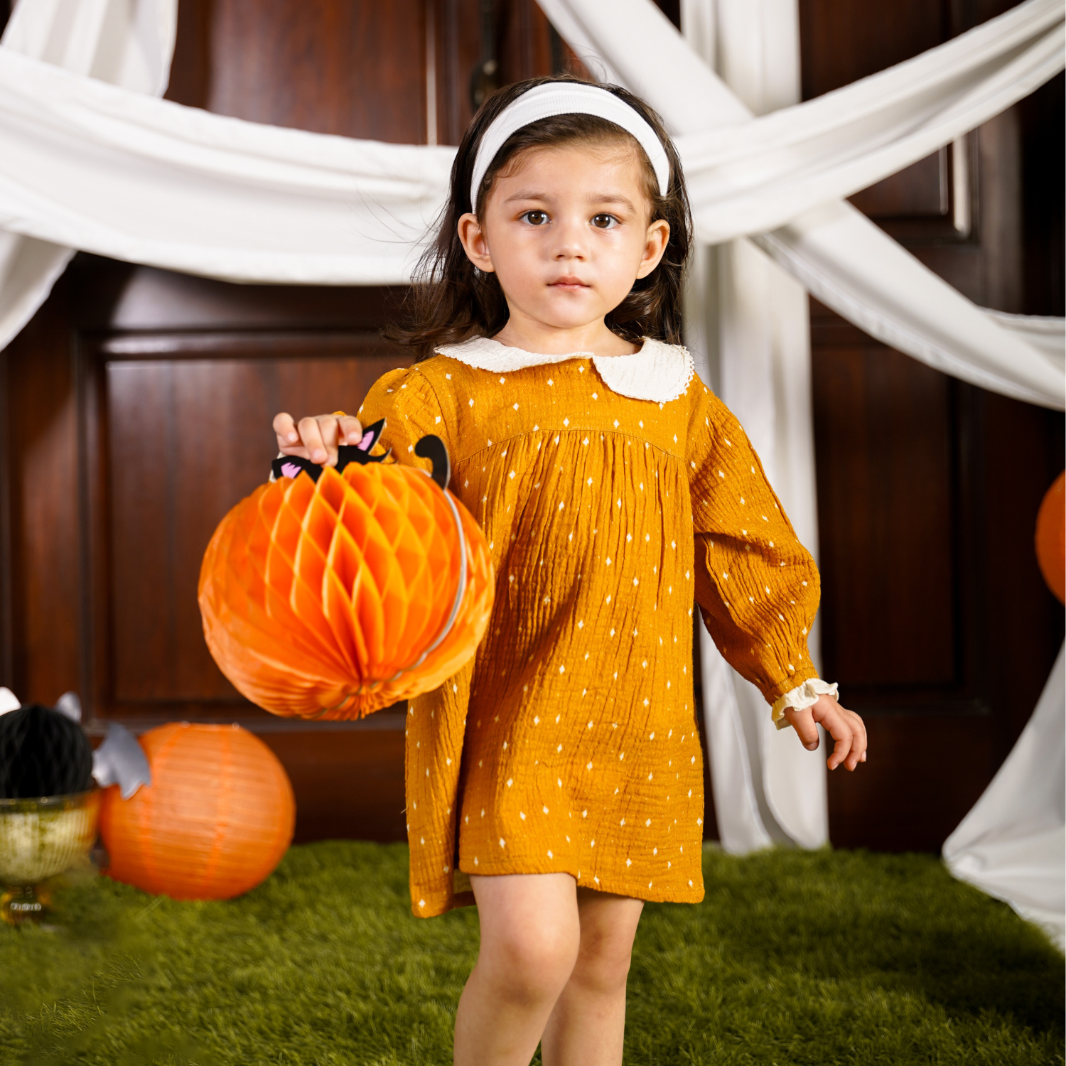 A baby girl wearing a golden-yellow frock with white polka dots, featuring a lace-collared neckline and long puffed sleeves. Perfect for a child's wardrobe.
