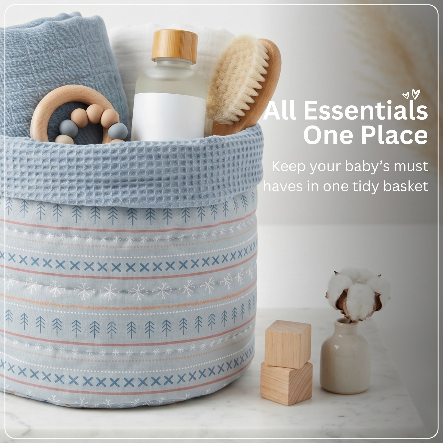 A light blue waffle-weave baby basket with Nordic patterned stripes, filled with baby essentials including a wooden teething ring, a bottle, a brush, and a folded cloth. The basket sits on a marble surface next to small wooden blocks and a small vase with cotton, with text overlay reading 'All Essentials One Place'.