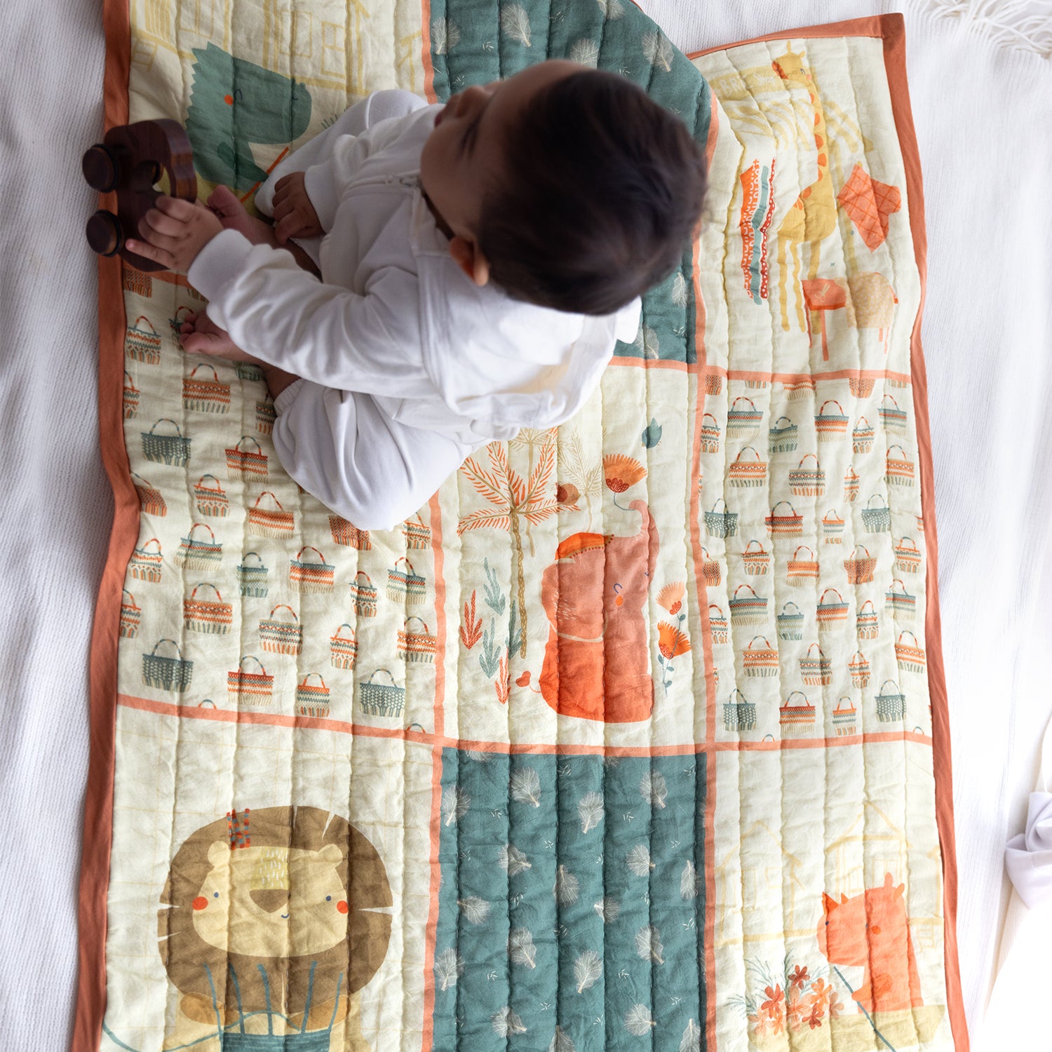 A baby in a white onesie sits on a patterned baby quilt, playfully holding a wooden toy and reaching for the colorful design.