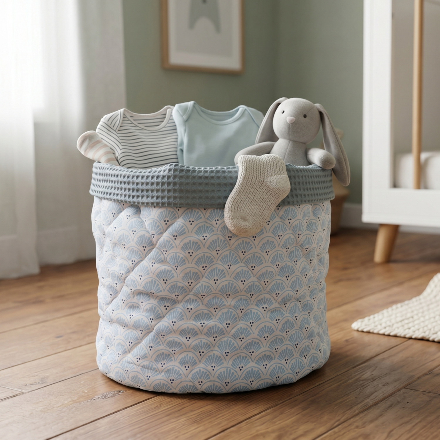 A light blue and white patterned fabric storage basket with a gray waffle-trimmed rim, styled with baby clothes, a plush bunny toy, and a knit bootie in a softly lit nursery room.