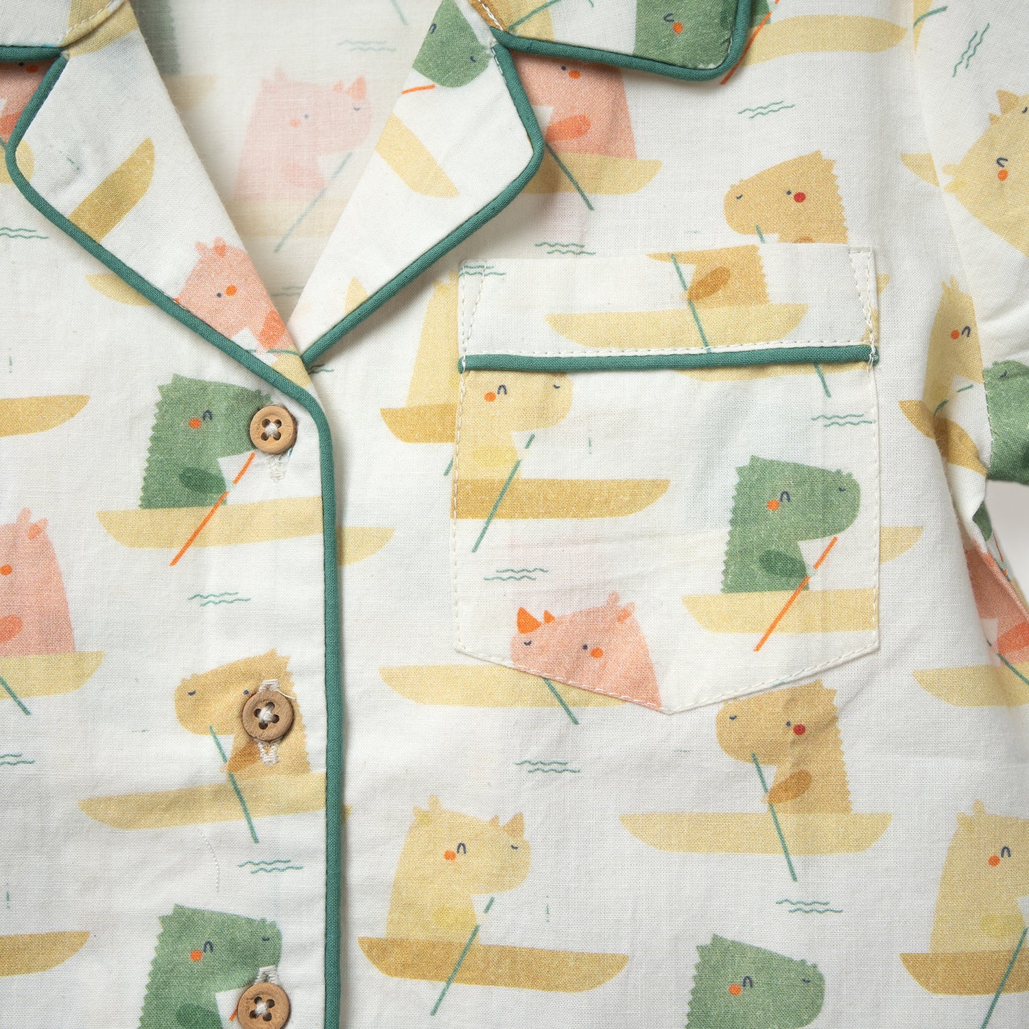 Closeup of a light-colored baby shirt featuring a playful print of animals in boats, bordered with green piping and wooden buttons.