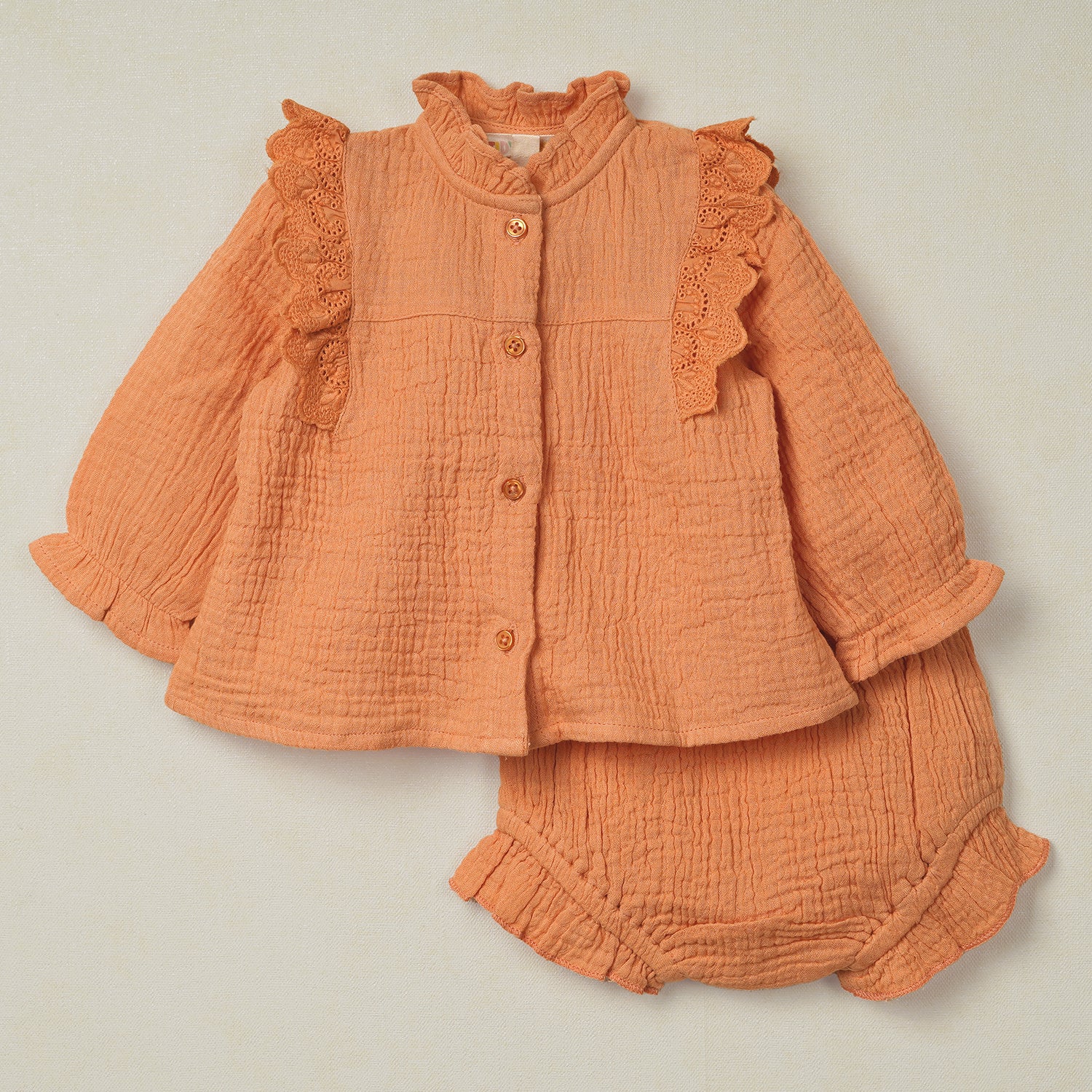 Adorable baby outfit featuring a textured muslin top with lace ruffle details and matching bloomers in a warm peach hue.
