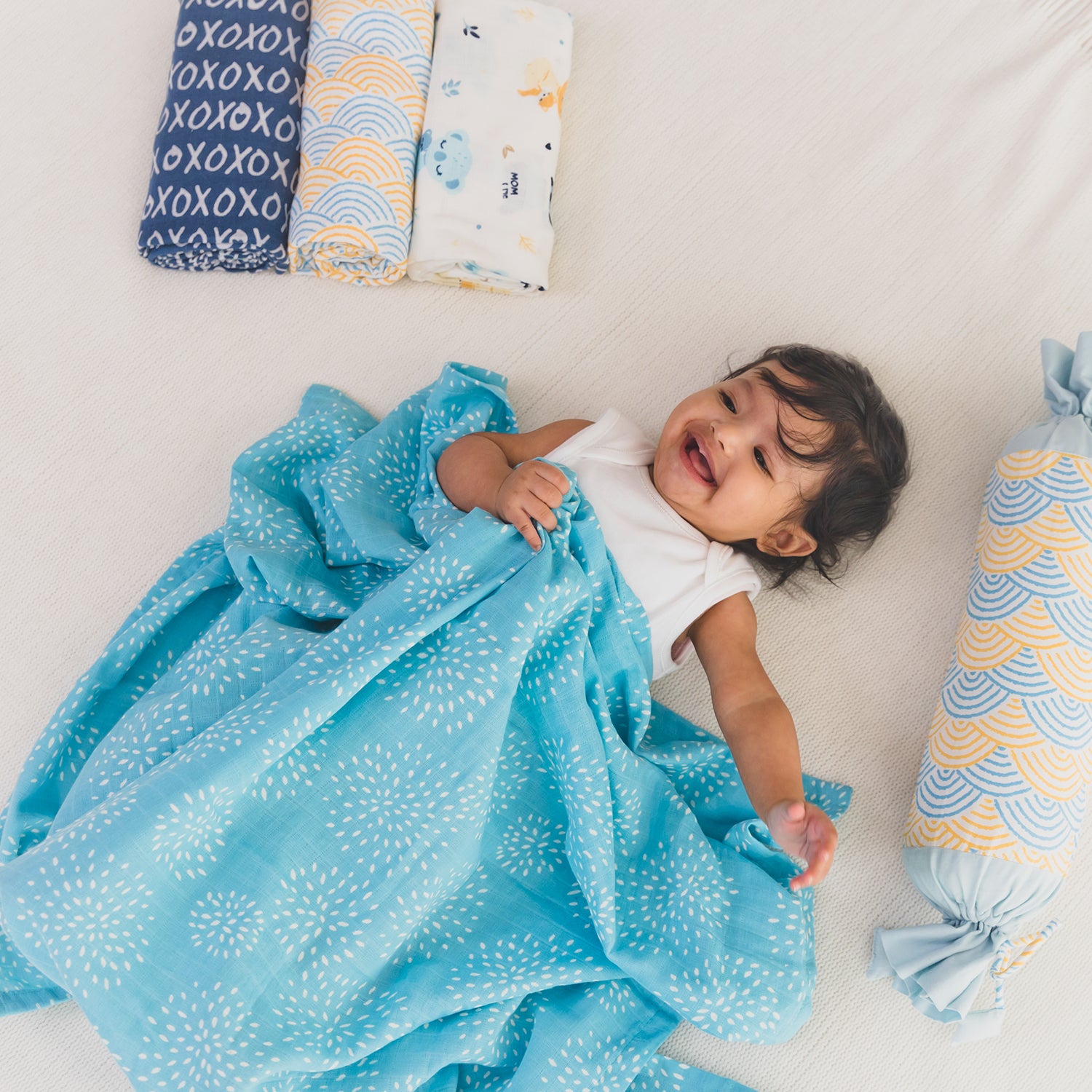 A smiling baby lying on a soft surface, wrapped in a vibrant blue blanket with white dotted patterns.