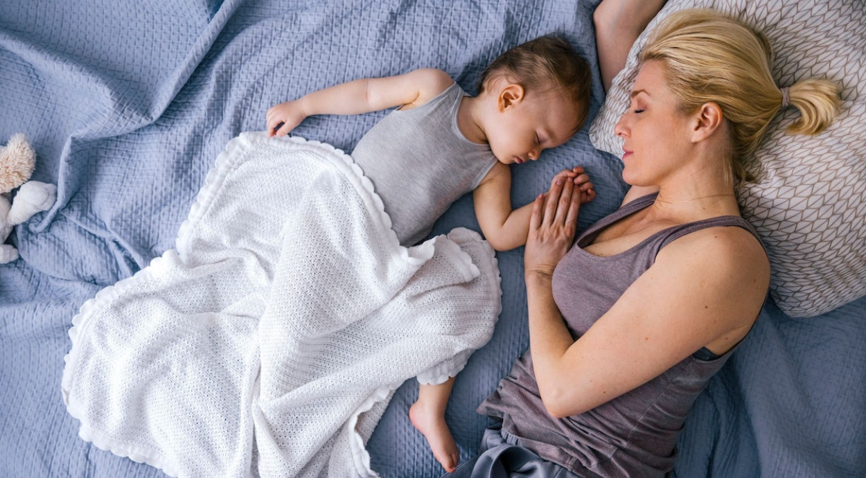 Mother and child lying on a bed, holding hands, with a white blanket partially covering the child on a blue quilted bedspread.