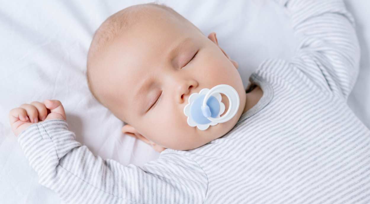 A baby lies on a soft white surface, wearing a gray and white striped onesie, with a pacifier in their mouth and their arms stretched out.