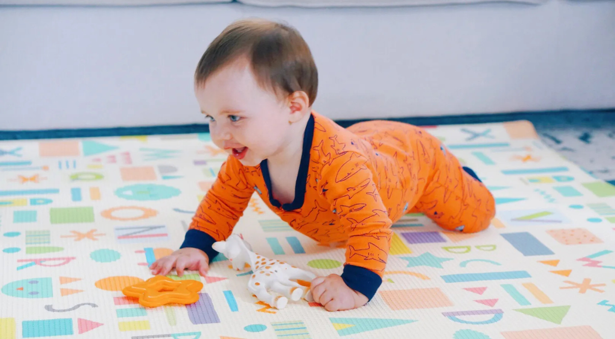 Baby in orange pajamas crawling on a colorful alphabet play mat, holding a giraffe toy and an orange star-shaped toy.