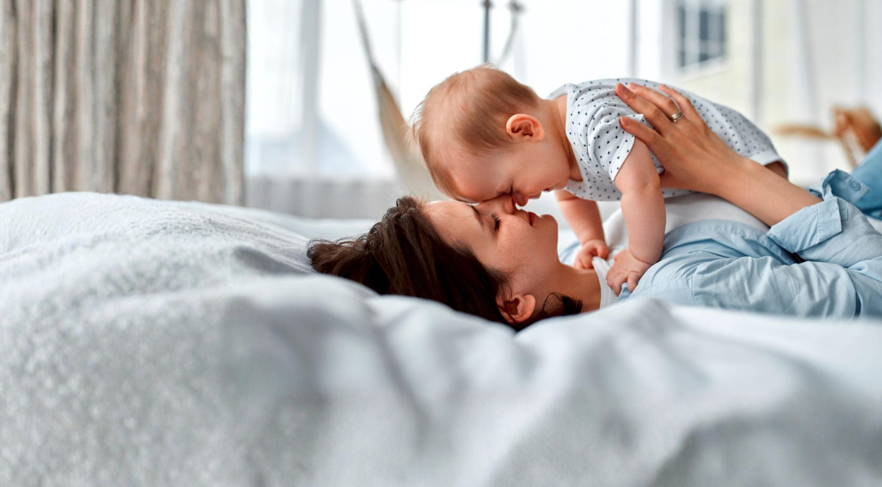 A mother lies on a bed, playfully lifting her baby above her, surrounded by soft bedding and warm natural light.