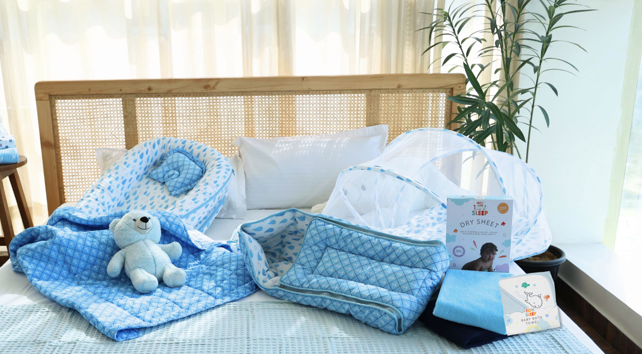A cozy bed setup featuring a blue and white baby bedding set, including a quilt, pillow, and canopy, with a blue teddy bear and baby book nearby. Bright, serene mood.