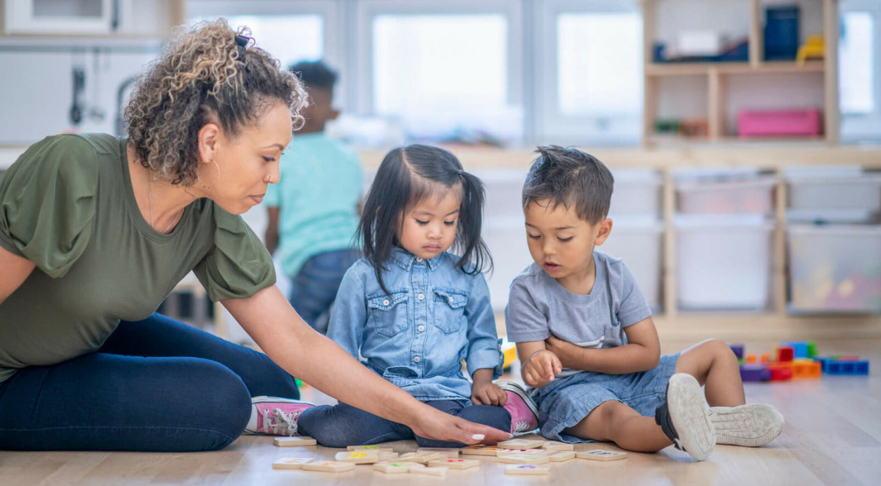 An adult interacts with two children on a play mat, engaging them with wooden educational toys in a bright, playful environment.