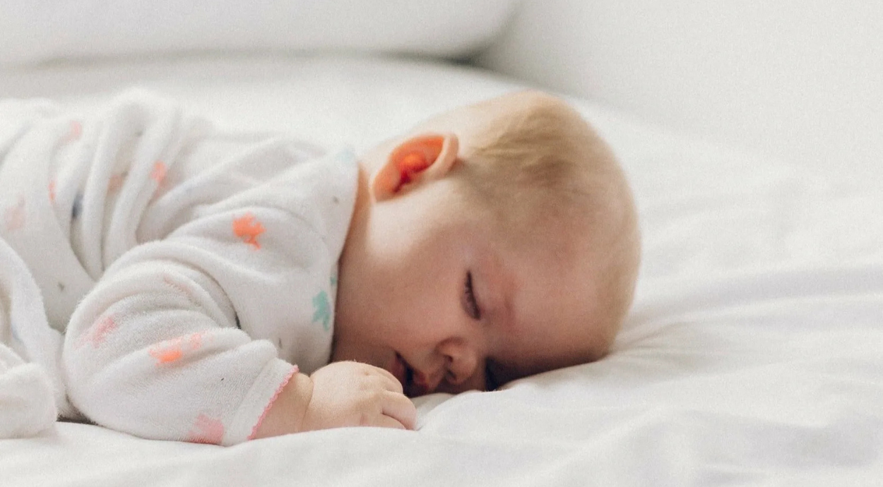 A baby in a colorful onesie is peacefully sleeping on a soft white bedspread.